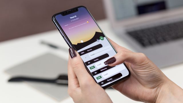 Hände halten ein Smartphone mit geöffneter Planungs-App, im Hintergrund ein Laptop