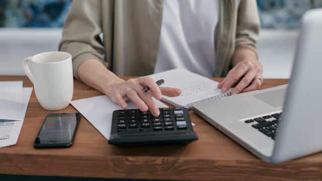Person kalkuliert am Schreibtisch mit Taschenrechner, Notizbuch und Laptop